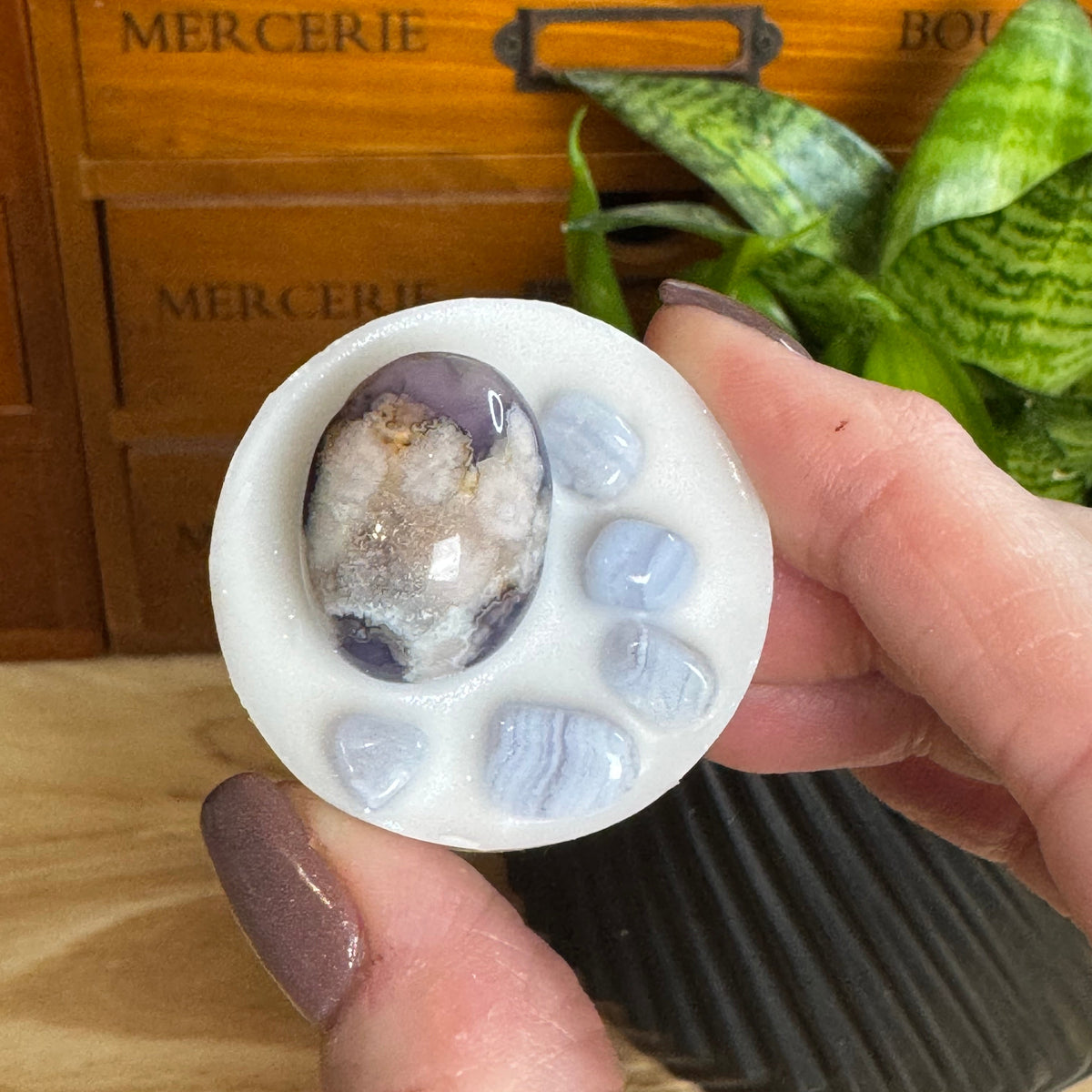 A hand holds a white circular stone holder featuring a polished purple and white agate cabochon in the center, surrounded by five small light blue baroque stones. The background shows a wooden drawer with 'MERCERIE' text and a green plant.