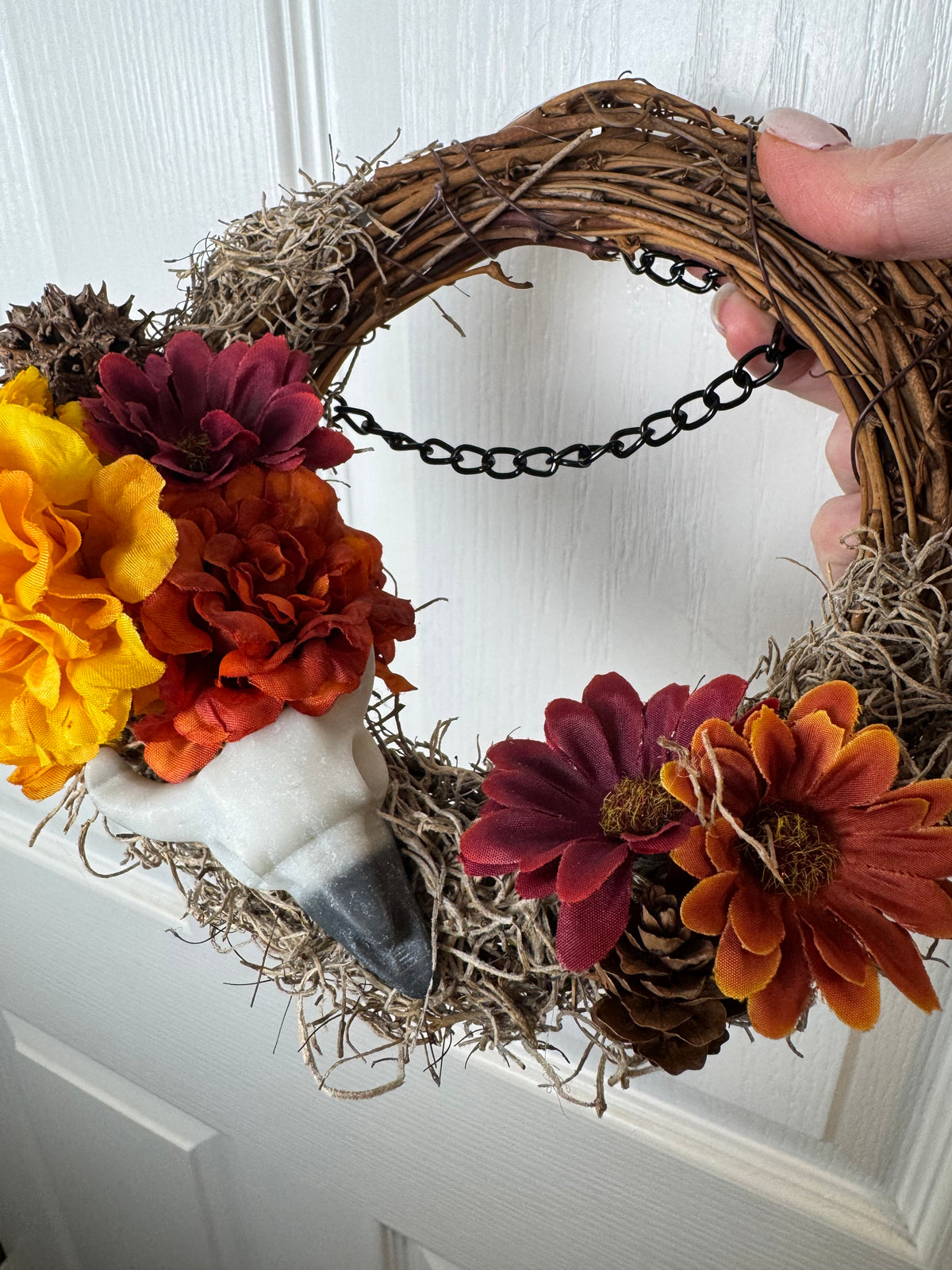 A grapevine autumn wreath with faux flowers in yellow, orange, and burgundy, a white bird figurine, and pinecones, held by a hand against a white paneled door.
