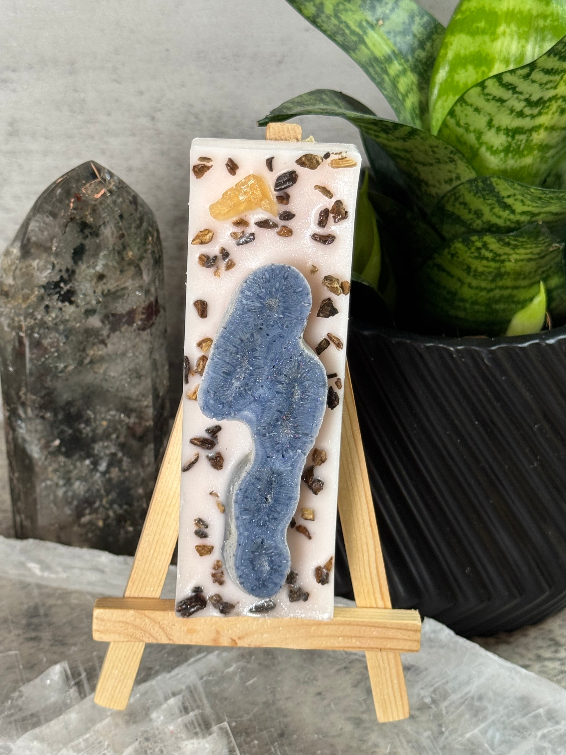 Decorative soap bar with natural elements on a small wooden stand, placed on a textured surface with a plant in the background.