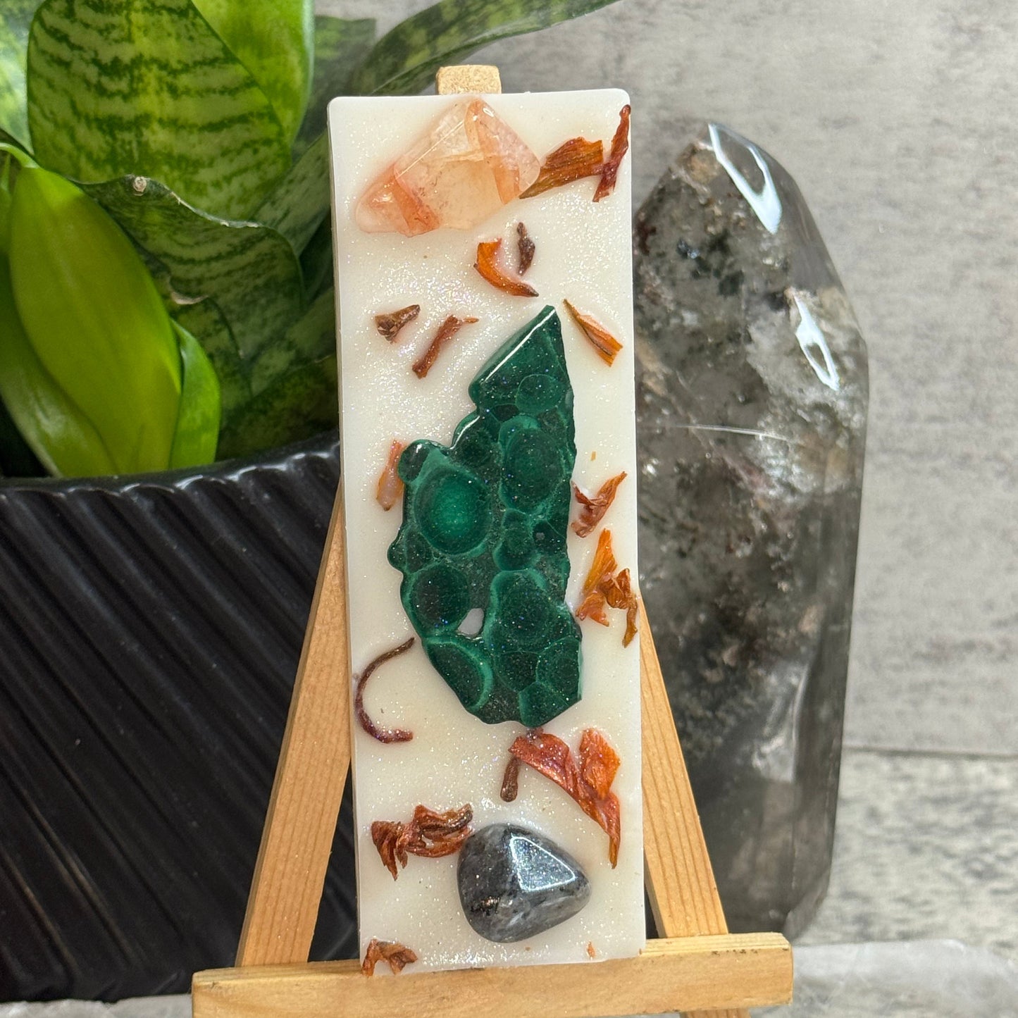 A rectangular cream-colored resin candle displayed on a small wooden easel. The candle features embedded natural stones, including a prominent green malachite piece, a peach crystal, and dried orange botanical pieces, set against a background with a snake plant and a clear quartz crystal.