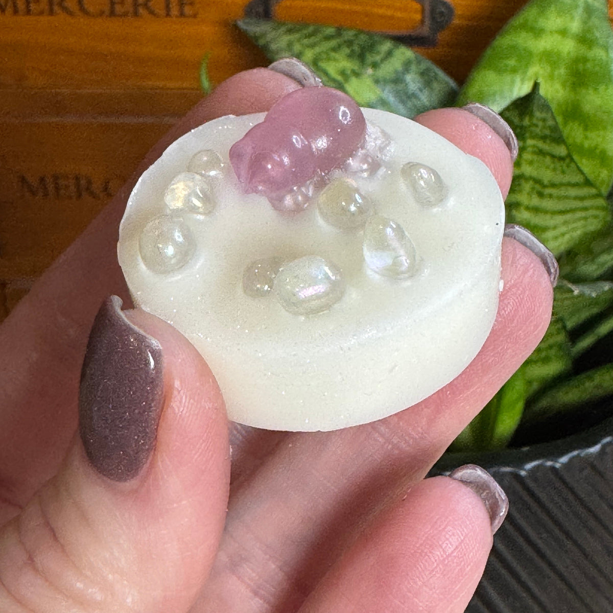 A hand with glittery mauve nail polish holds a round, off-white resin or wax melt featuring a small pink crystal and clear bubble details, set against a background of a wooden drawer and green snake plant leaves.