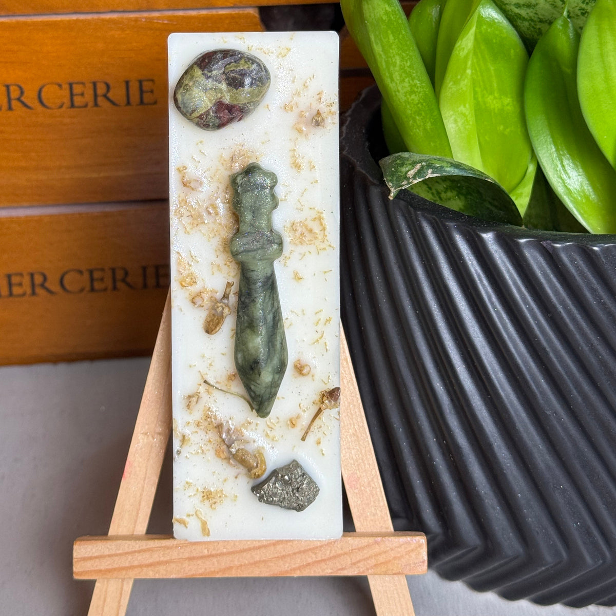 A vertical white stone plaque featuring a green stone figure and natural inclusions, displayed on a small wooden easel. The background includes a wooden drawer with 'CERIE' text and a potted green plant in a black ribbed pot.
