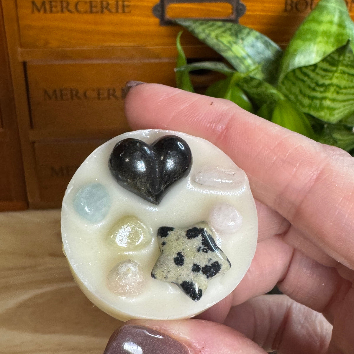 A hand holds a round cream-colored wax melt or soap with embedded polished stones, including a black heart, a black-and-white spotted star, and pastel-colored oval stones. The background shows a wooden drawer with 'MERCERIE' text and a green plant.