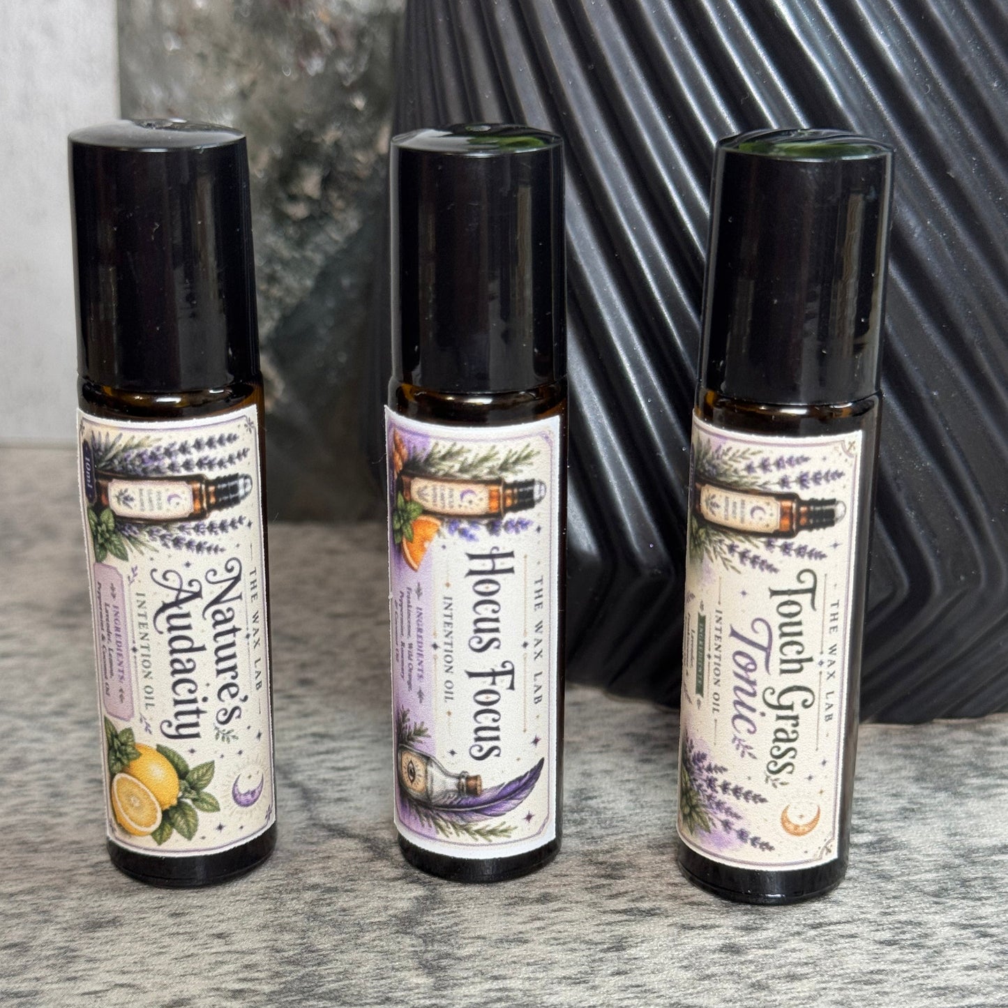 Three small roll-on bottles of essential oils from the brand 'The Alchemist' are lined up on a gray textured surface. The bottles are labeled 'Nature's Aquacade', 'Hocus Focus', and 'Touch Grass'.