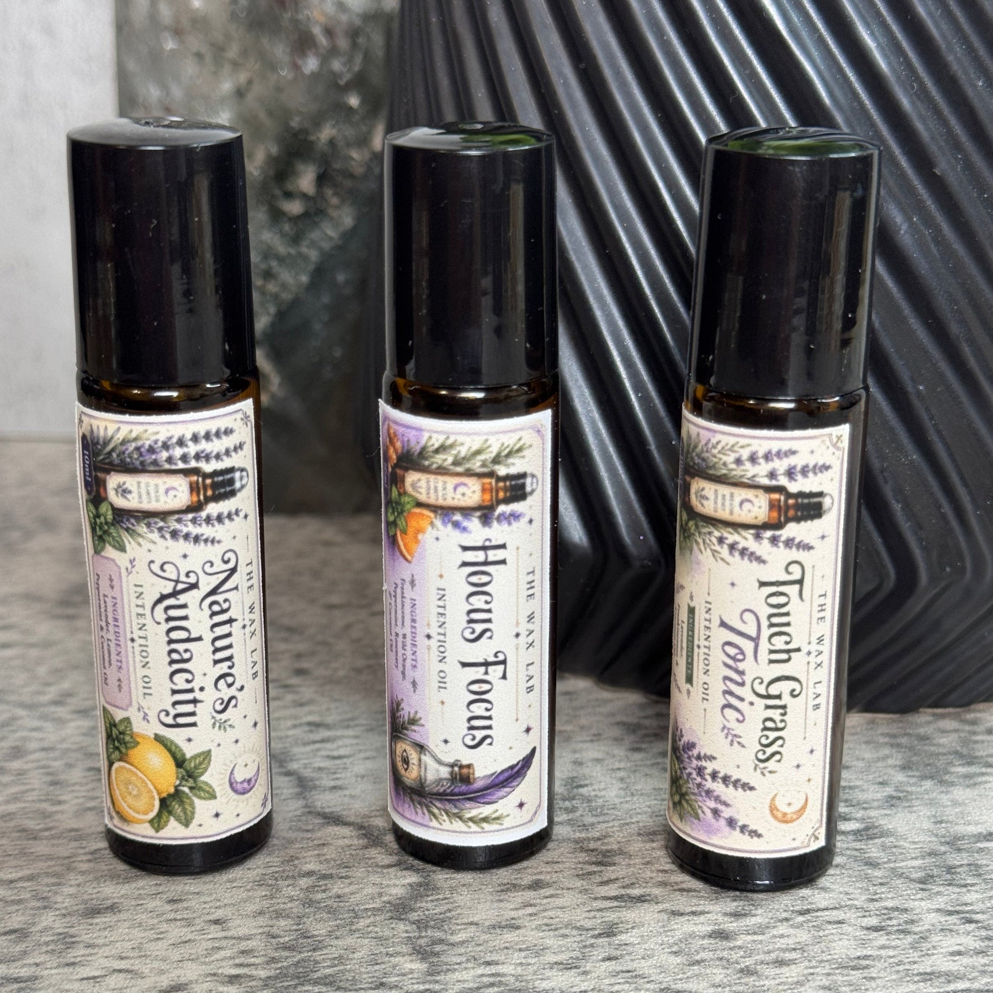 Three small roll-on bottles of essential oils from the brand 'The Alchemist' are lined up on a gray textured surface. The bottles are labeled 'Nature's Aquacade', 'Hocus Focus', and 'Touch Grass'.