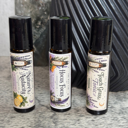 Three small roll-on bottles of essential oils from the brand 'The Alchemist' are lined up on a gray textured surface. The bottles are labeled 'Nature's Aquacade', 'Hocus Focus', and 'Touch Grass'.
