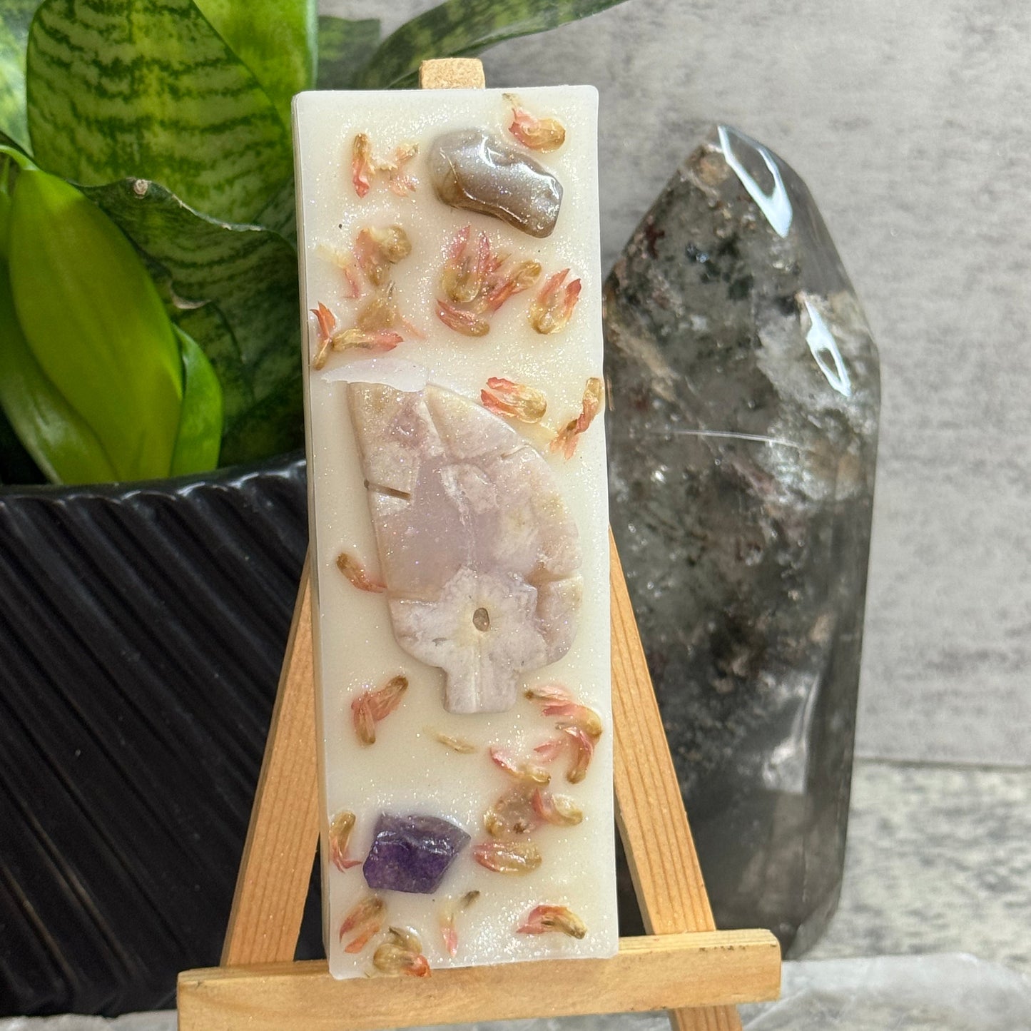 A rectangular handmade soap bar displayed on a small wooden easel. The cream-colored soap features embedded natural stones and dried botanicals, including a prominent pale pinkish-beige stone in the center and a purple crystal near the bottom.