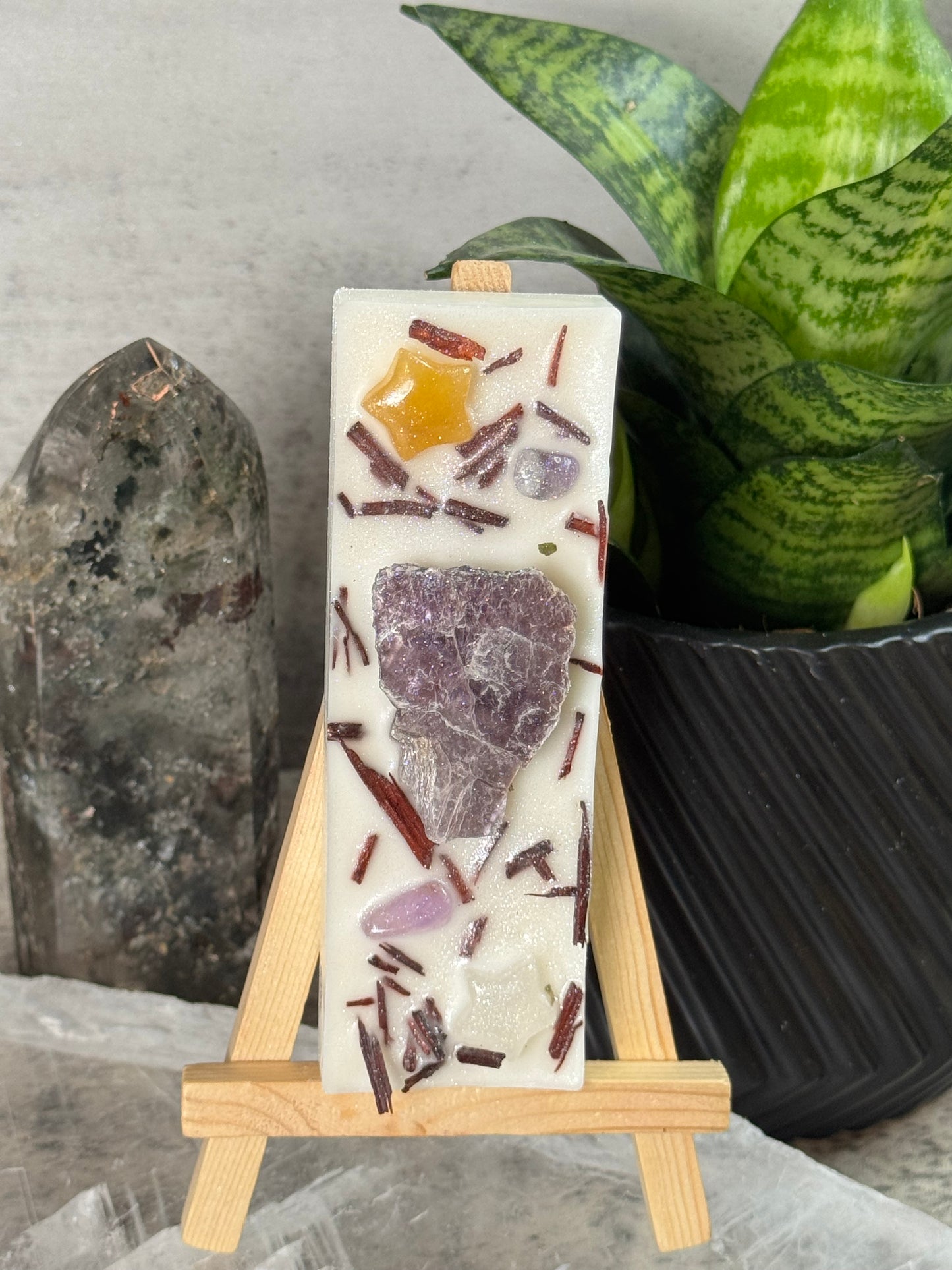 Decorative soap bar with embedded stones on a wooden stand, placed on a stone surface with a plant in the background.