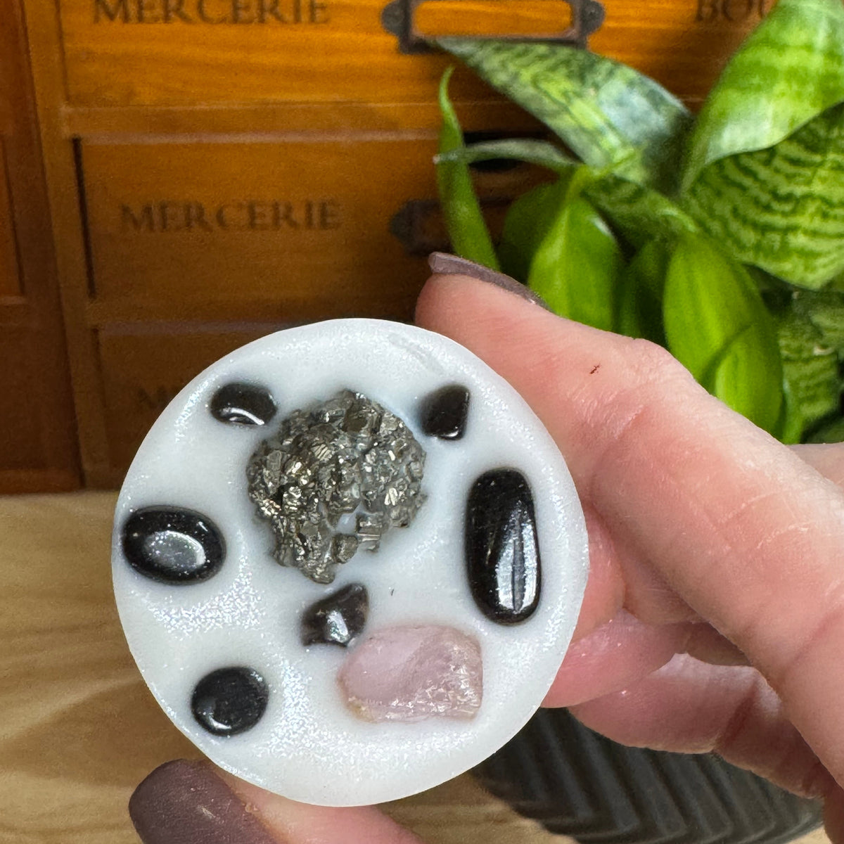 A hand holds a round white resin coaster featuring a central silver-gray crystal cluster, surrounded by black and pink decorative stones, with a wooden drawer and green plant in the background.