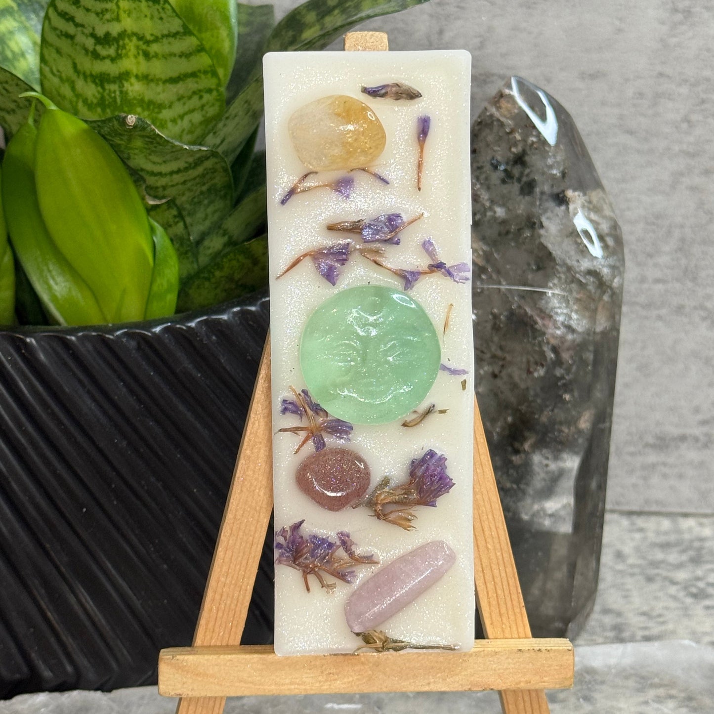 A rectangular cream-colored resin candle displayed on a small wooden easel. The candle features embedded polished crystals in amber, green, pink, and brown, along with dried purple flowers.
