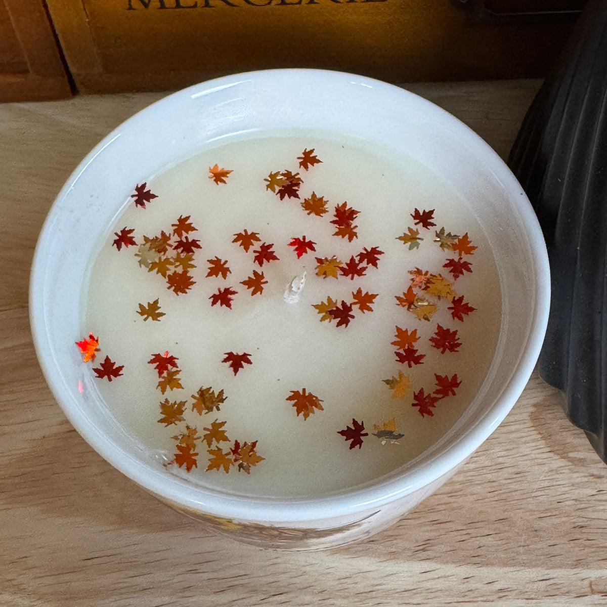 A white ceramic bowl filled with a creamy off-white wax or soap, decorated with scattered dried red, orange, and yellow maple leaves, set on a light wood surface with a wooden box in the background.