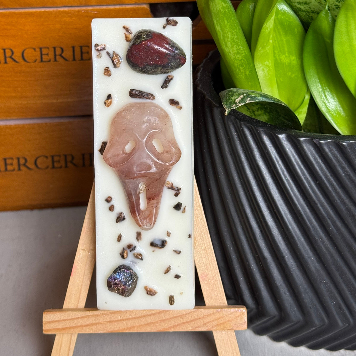 A rectangular white resin candle displayed on a small wooden easel, featuring a carved pink skull, dark brown stones, and scattered chocolate pieces. The background includes a wooden drawer and a green potted plant.