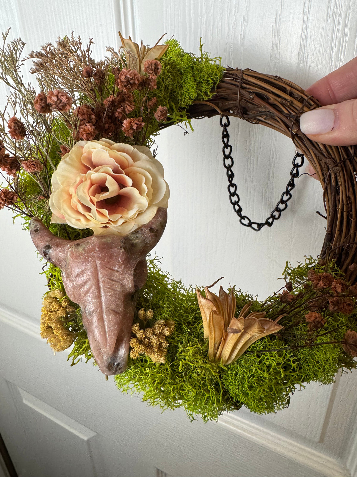 A rustic grapevine wreath decorated with green moss, dried brown flowers, a large pale peach rose, and a realistic pinkish-brown skull, being held against a white paneled door. A person's hand is visible on the right side.