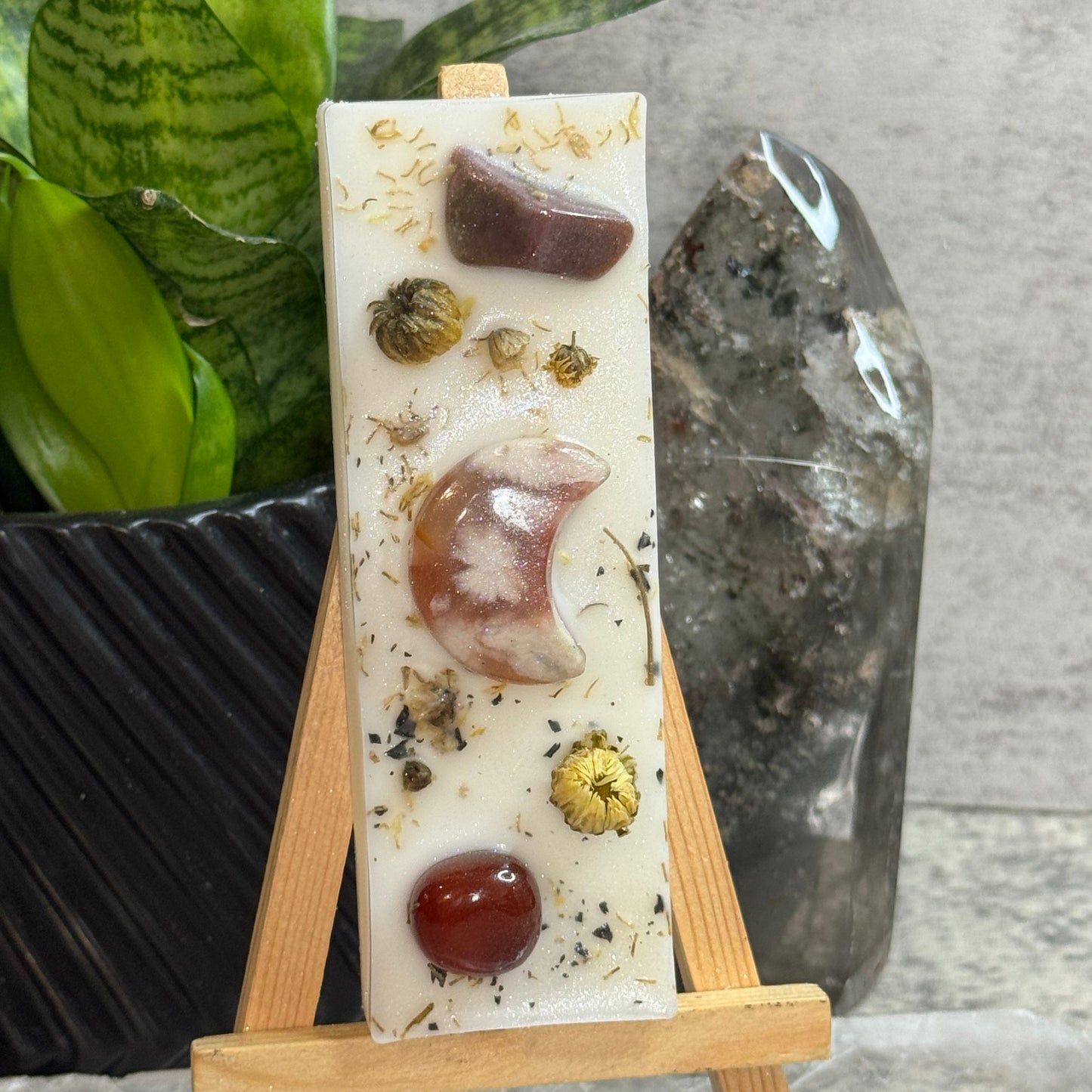 A rectangular white resin candle displayed on a small wooden easel. The candle contains embedded natural elements including dried flowers, a crescent moon, a dark brown stone, and a red gem.
