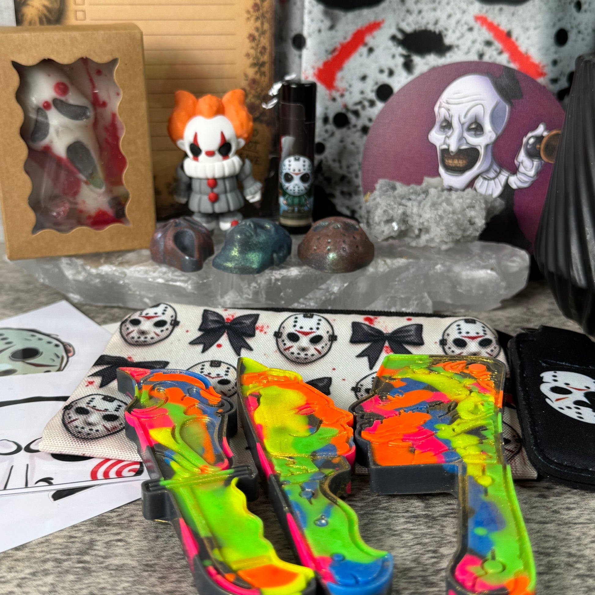 A collection of gothic and horror-themed items laid out on a textured gray floor, including neon tie-dye high heels, a Pennywise collectible figure, a skull-patterned bag, and other spooky accessories.