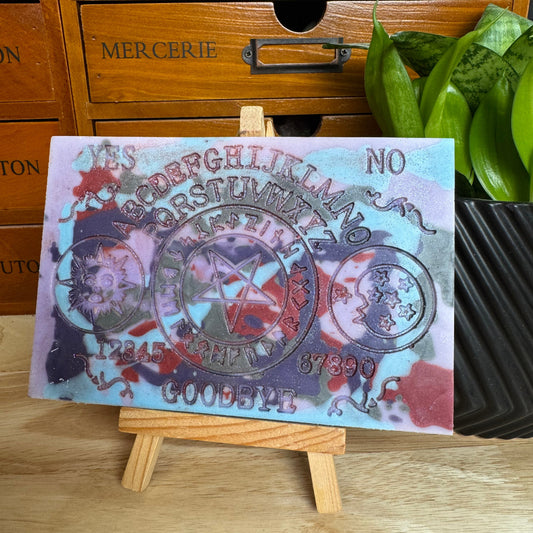 A rectangular tarot card art print featuring a pentagram and occult text, displayed on a small wooden easel in front of a wooden drawer cabinet and a green plant.