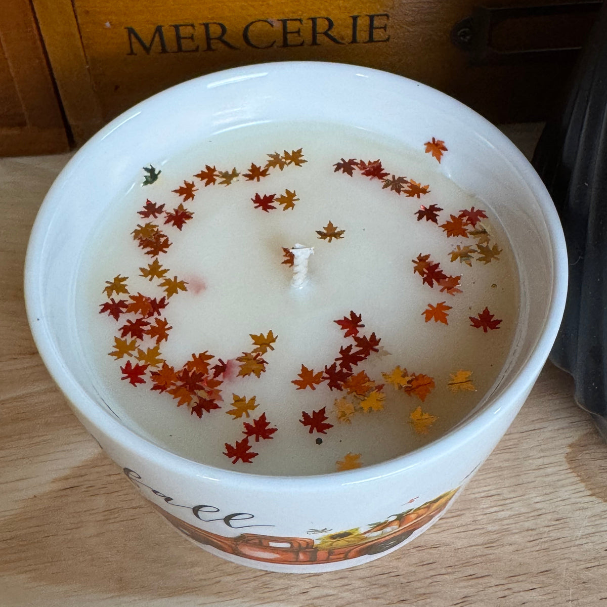 A white ceramic candle jar with a single wick, filled with creamy off-white wax and decorated with small dried orange, red, and yellow maple leaves floating on the surface. The jar is placed on a light wooden table, with a wooden drawer labeled 'MERCERIE' in the background.