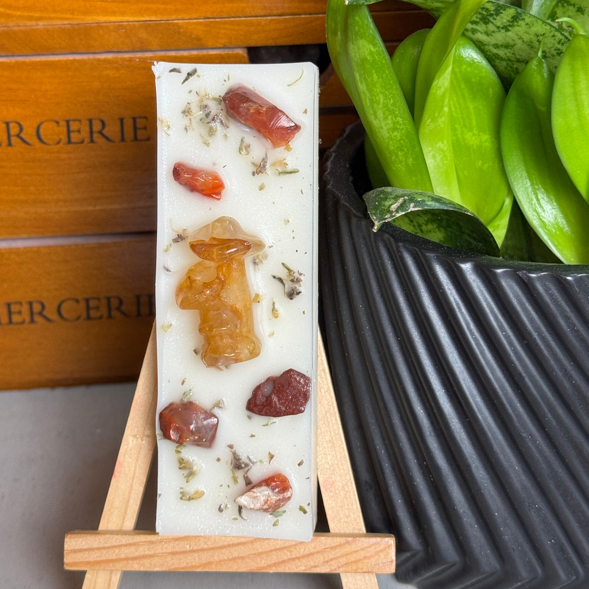 A rectangular white soap bar with embedded amber and red stones, displayed on a small wooden easel. The background features a wooden drawer with 'BUCERIE' text and a potted green plant in a black ribbed pot.