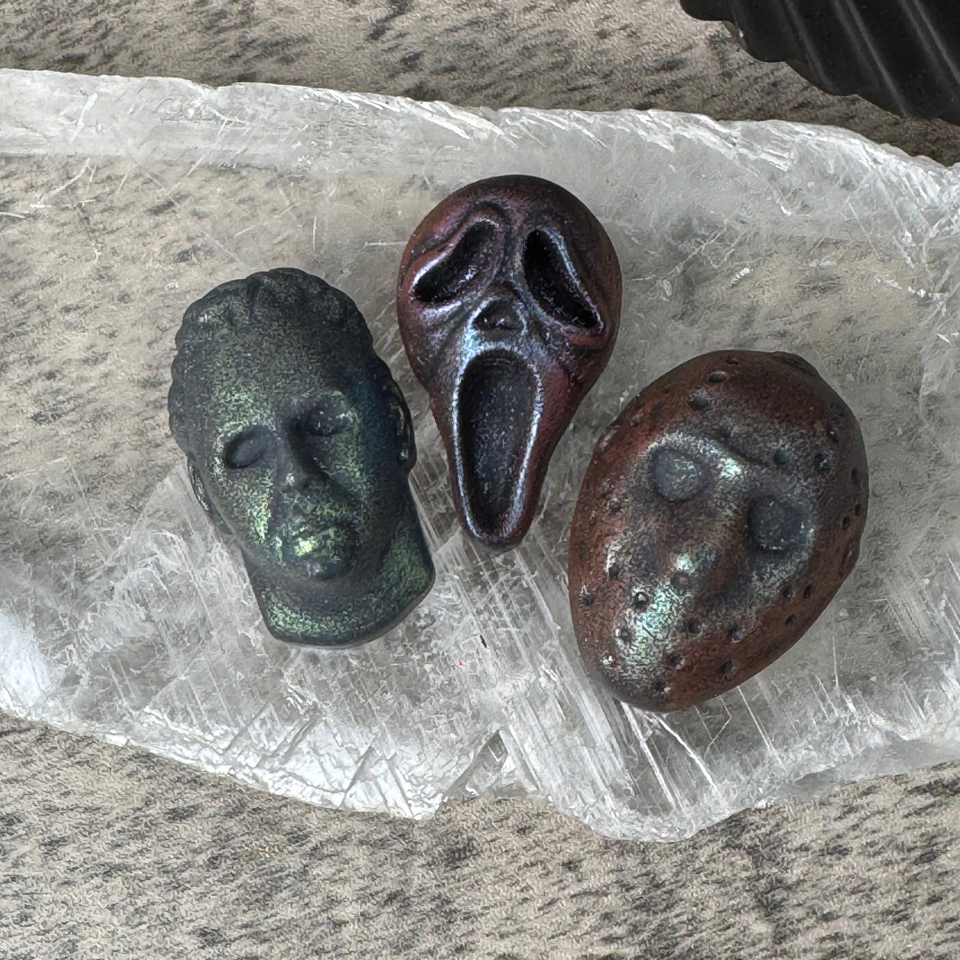 Three small decorative masks or sculptural objects in dark metallic and earthy tones, arranged on a crumpled clear plastic sheet over a speckled gray surface.
