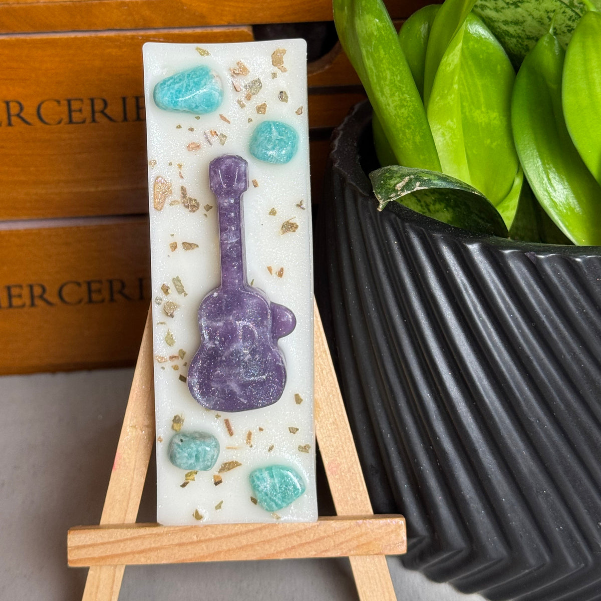 A rectangular white resin plaque featuring a purple guitar design and turquoise stones, displayed on a small wooden easel. The background features a wooden drawer and a potted green plant.