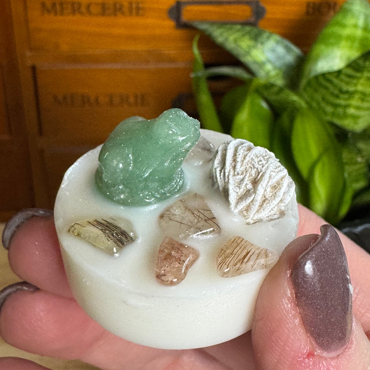 A hand with dark brown manicured nails holds a small white circular soap or candle featuring a green frog topper, a white textured crystal cluster, and several small natural mineral chips. The background shows a wooden drawer with engraved labels and green plants.