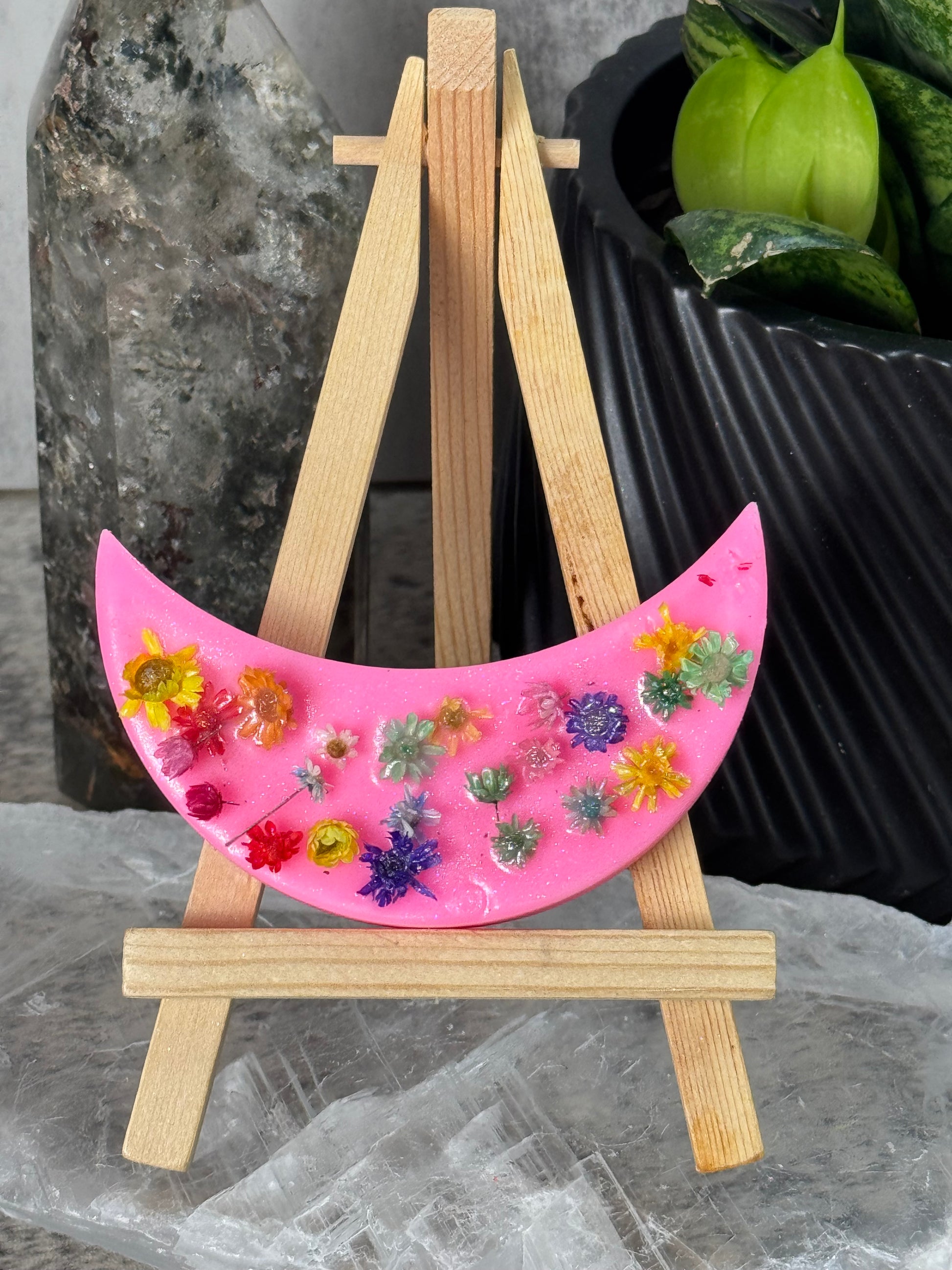Pink crescent moon-shaped object with floral designs on a wooden stand against a dark background.