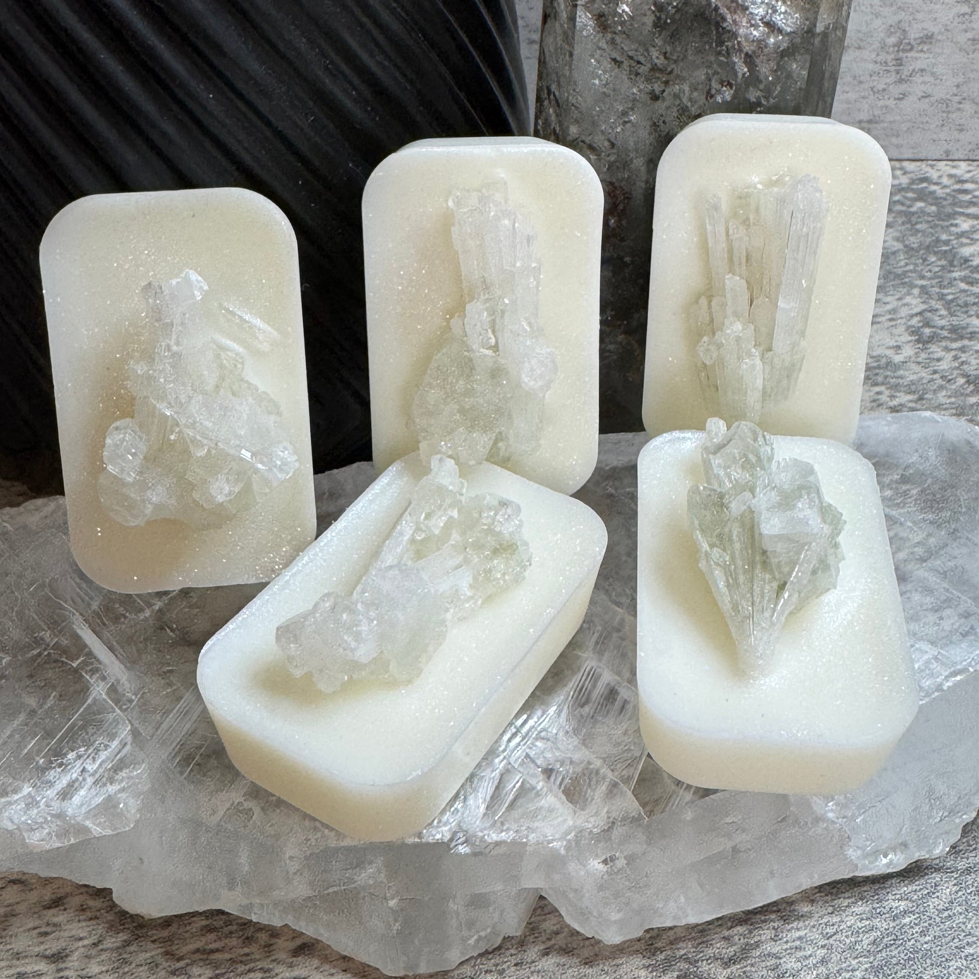 Four rectangular cream-colored soap bars, each topped with a clear crystal cluster, arranged on a large clear quartz crystal with a dark ribbed vase and gray stone in the background.
