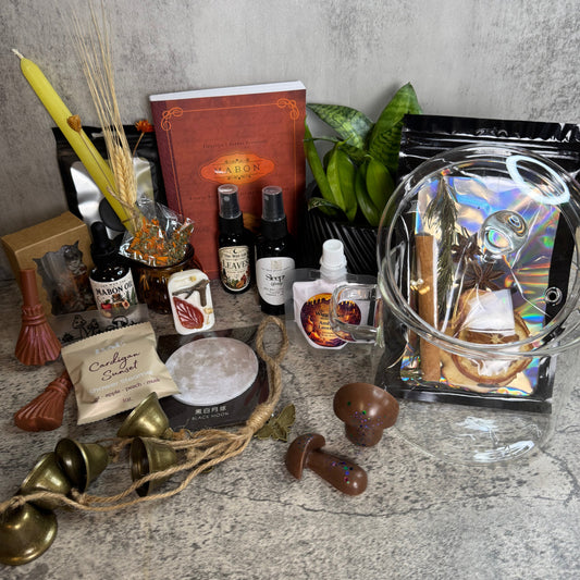 A styled flat-lay of a ritual or spiritual kit on a gray stone surface against a textured gray wall. The kit includes a red book, two small bottles, a pouch labeled 'Cowboy Candles', a clear plastic bag with an iridescent insert, brass bells, wooden and metallic mushroom-shaped pieces, a candle, dried wheat, and other small accessories.