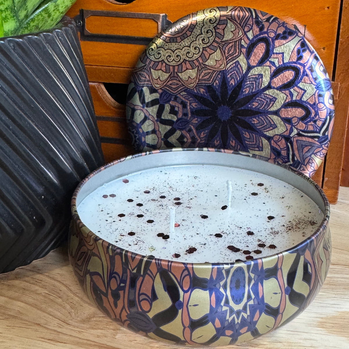 A round metal candle tin filled with white wax and topped with dark speckles, shown next to its matching lid on a wooden surface. The tin and lid feature an intricate mandala-style pattern in navy blue, gold, and brown.