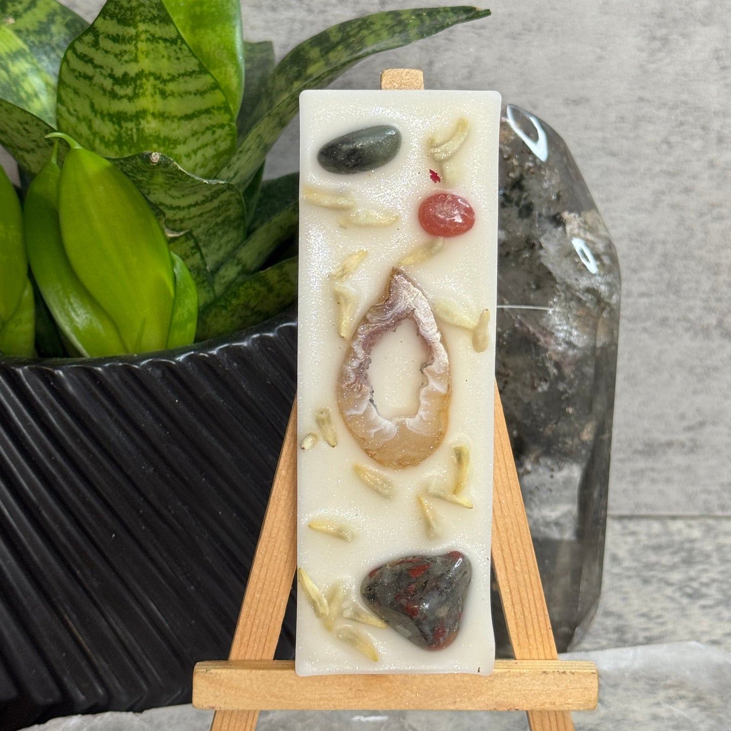 A rectangular cream-colored resin candle displayed on a small wooden easel. The candle features embedded natural stones, dried botanical pieces, and a central agate slice.