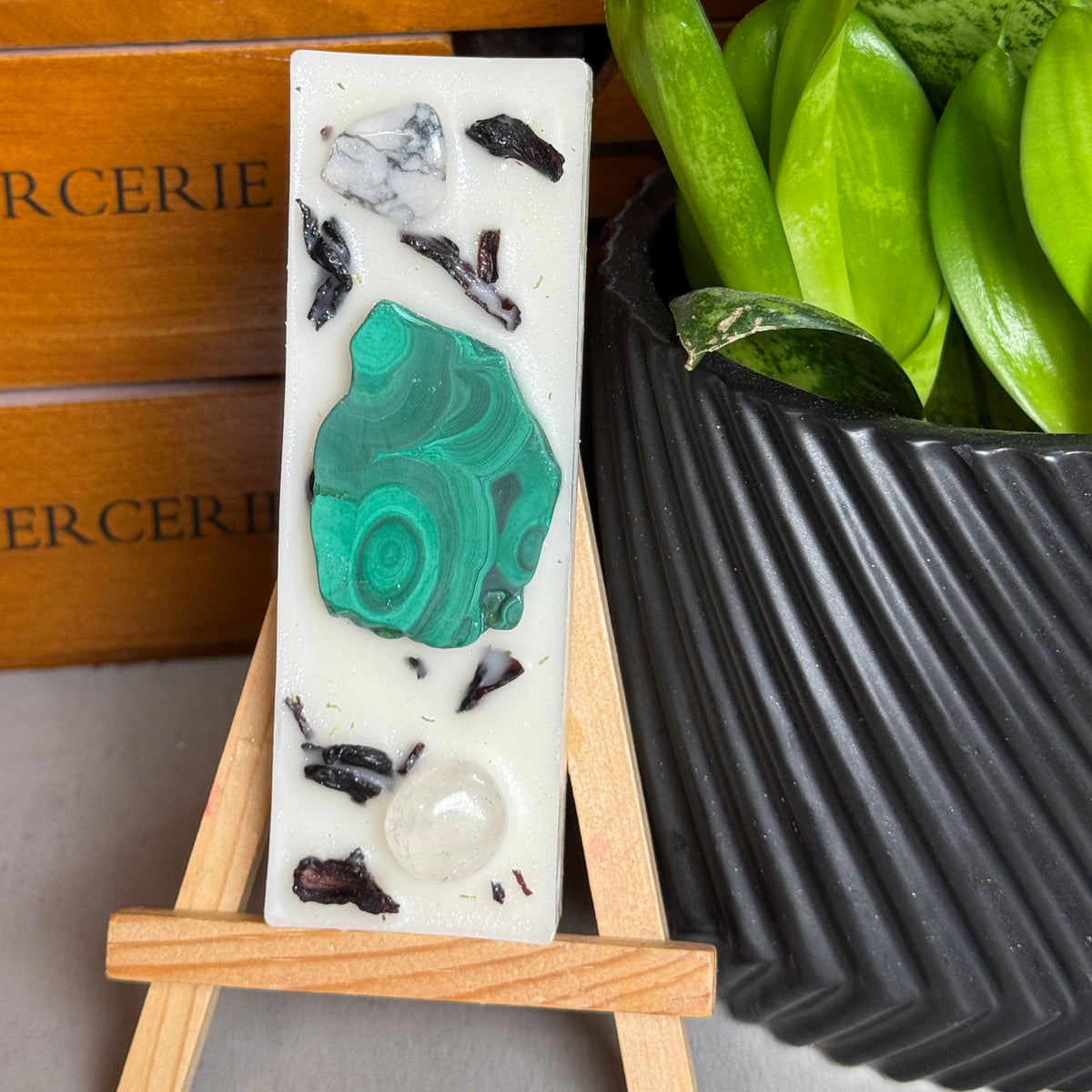 A rectangular white resin candle with a large green malachite stone and black/dark inclusions, displayed on a small wooden easel next to a potted green plant.