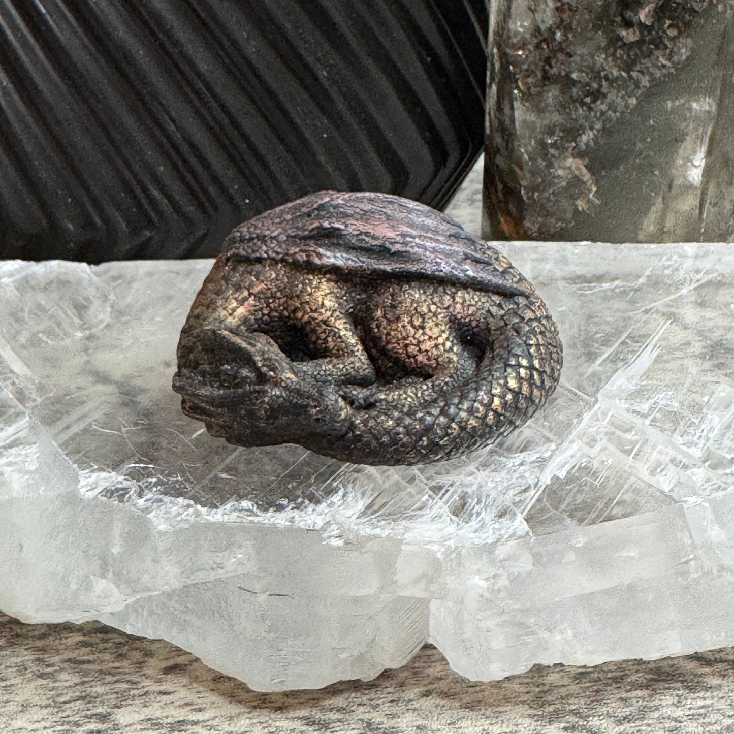 A small carved figurine of a sleeping dragon or lizard with textured scales, displayed on a clear crystal cluster with dark decorative objects in the background.