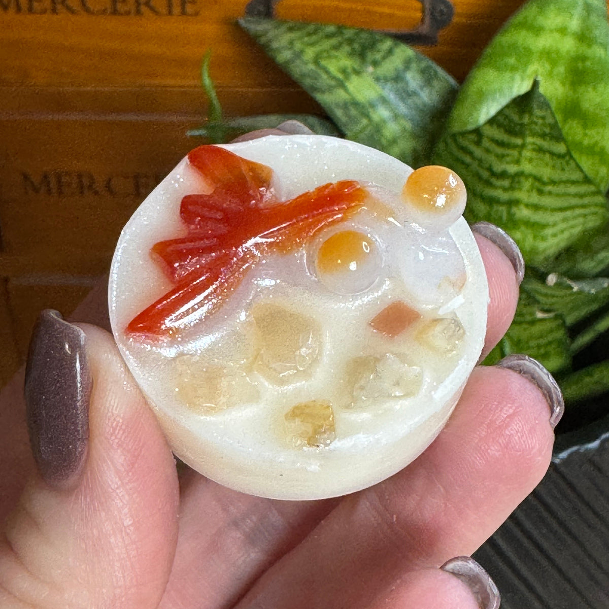 A hand with glittery mauve nails holds a small round carved stone pendant featuring a red fish and orange bubbles on a white background, with a wooden drawer and green plants in the background.