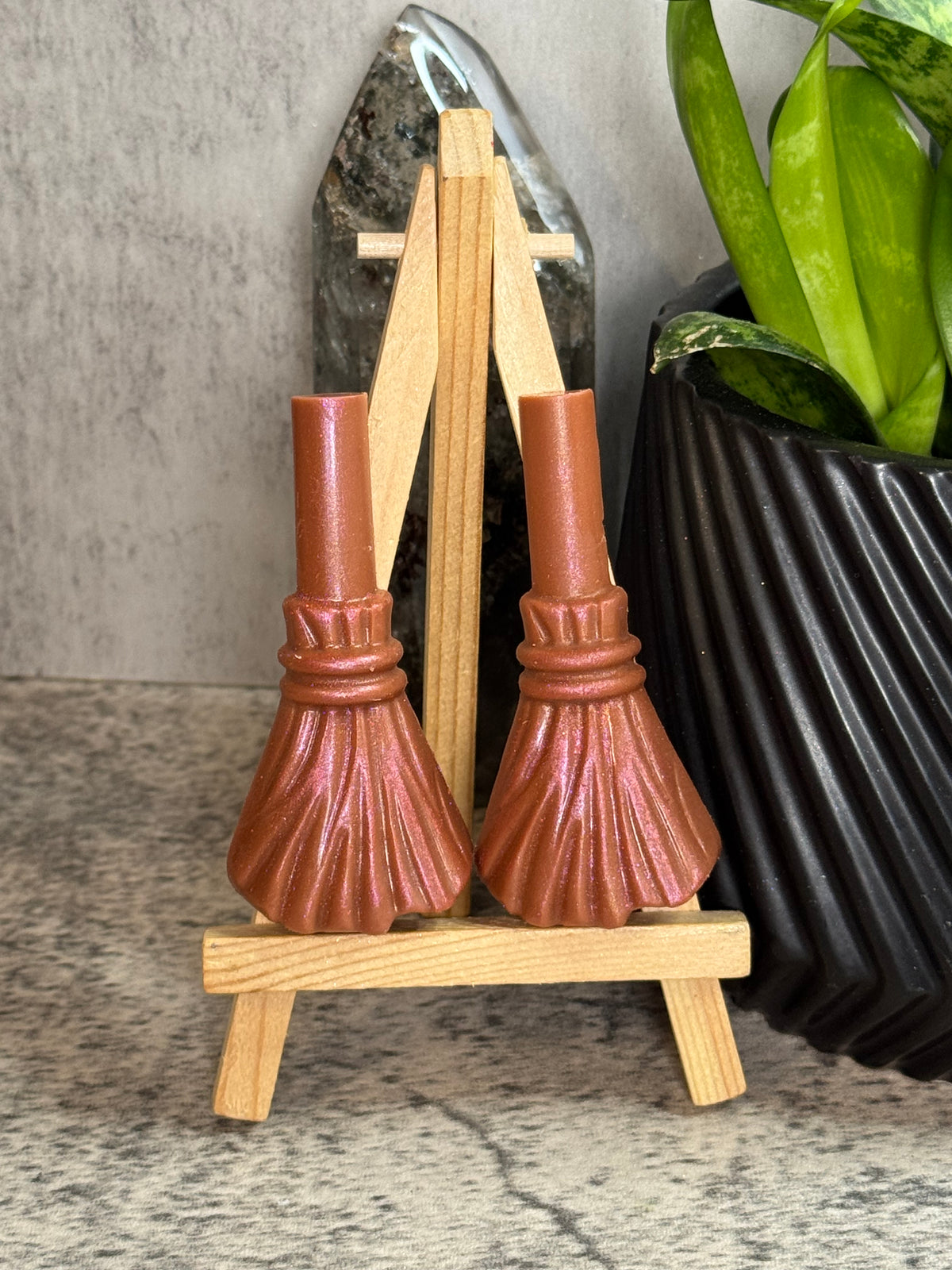 Two small witch broomstick figurines with metallic copper-brown handles and dark bristles, displayed on a miniature wooden easel stand.