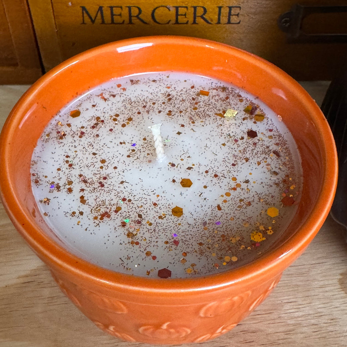 An orange ceramic candle bowl filled with white wax and decorated with multicolored glitter, set on a wooden surface with a wooden drawer in the background.