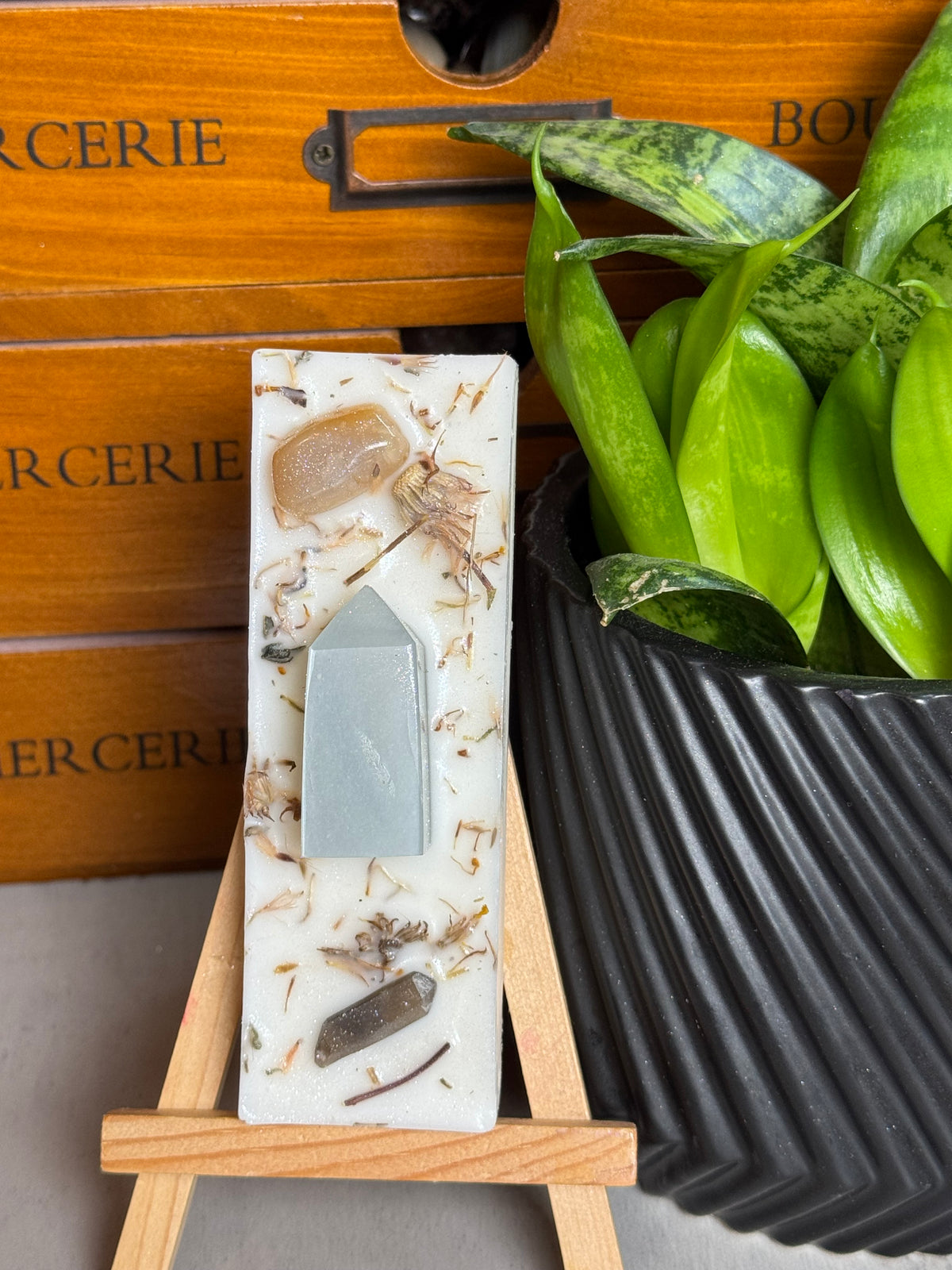 A rectangular white resin candle with embedded botanical pieces and crystals, displayed on a small wooden easel. The background features a wooden drawer with 'BUCERIE' text and a green potted plant in a black ribbed pot.