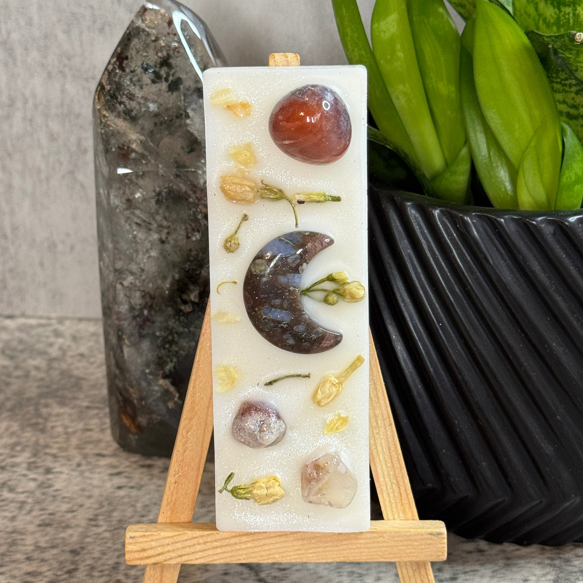A rectangular white resin candle displayed on a small wooden easel. The candle features embedded natural stones, dried flower petals, and a crescent moon charm.