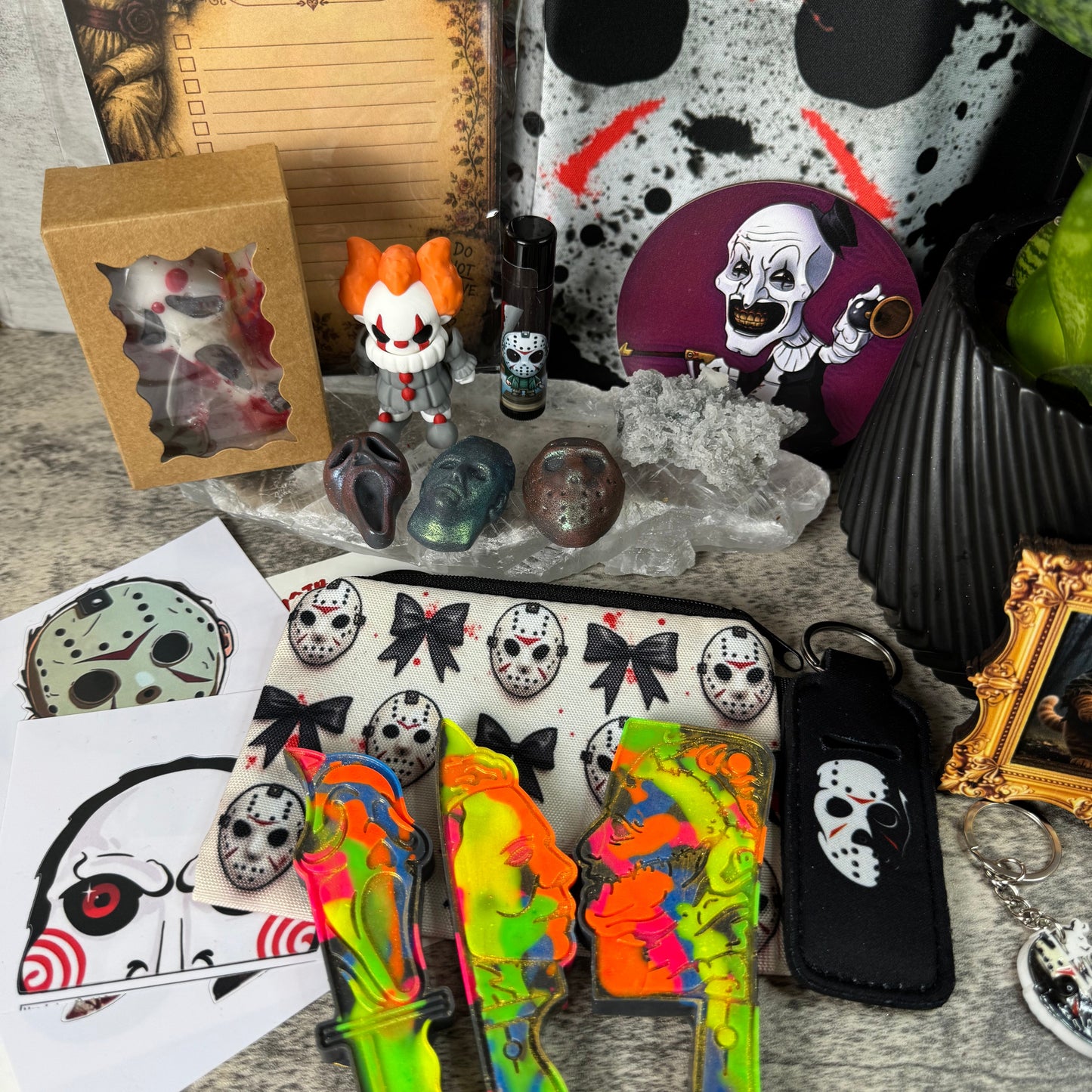 A collection of horror-themed merchandise arranged on a stone surface, including a zip pouch with repeated hockey mask graphics, two brightly colored tie-dye knife-shaped accessories, printed character art cards, a small figurine, a packaged item in a window box, and other spooky accessories.