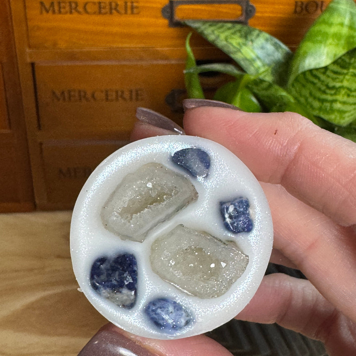 A hand holds a round white wax melt or soap featuring four geode-like crystal sections, including two pale gray crystals and two dark blue crystals. The background shows a wooden drawer with 'MERCERIE' text and a green plant.
