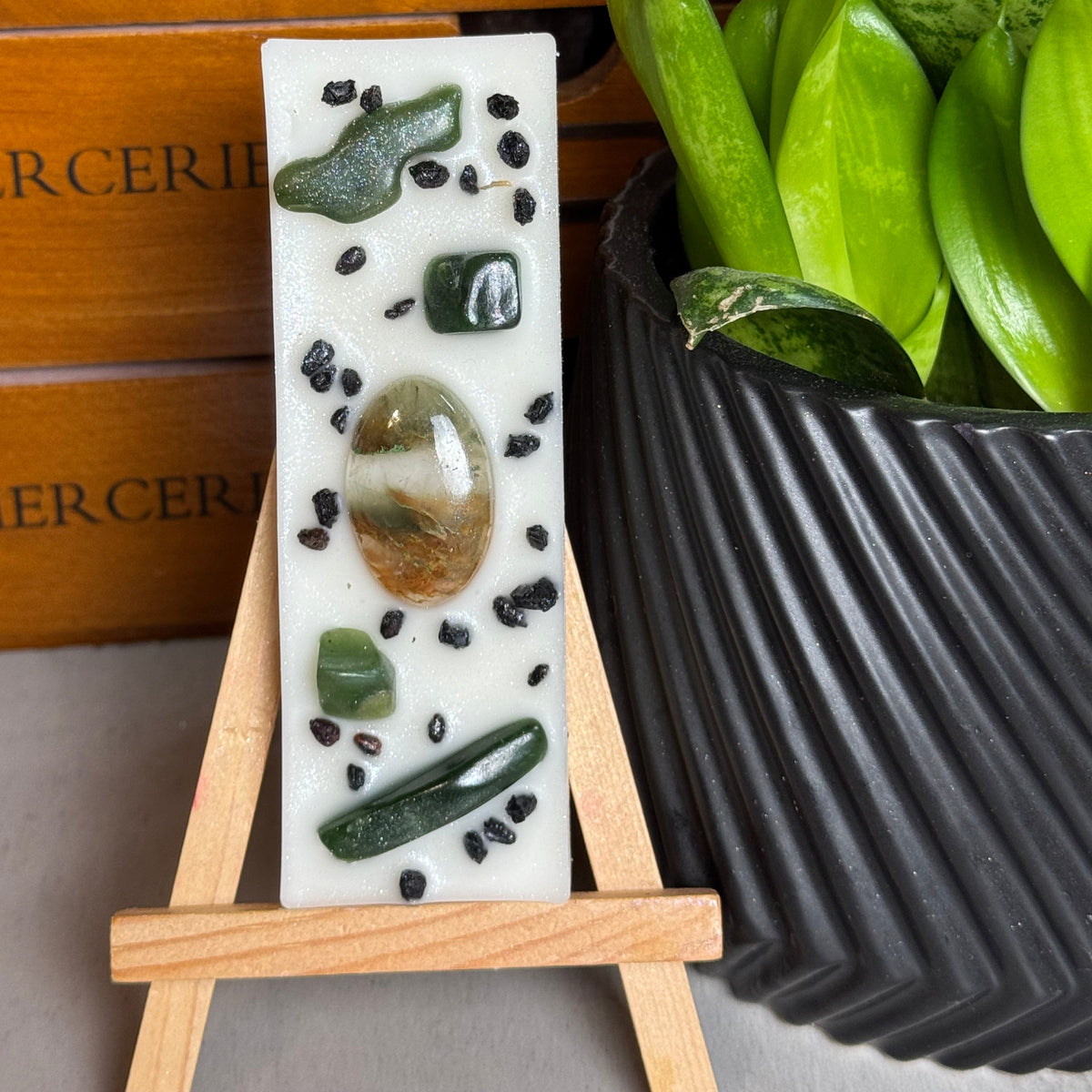 A rectangular white resin candle displayed on a small wooden easel, featuring embedded green crystals and black seed-like details. The candle is styled in front of a wooden drawer and next to a potted green plant.