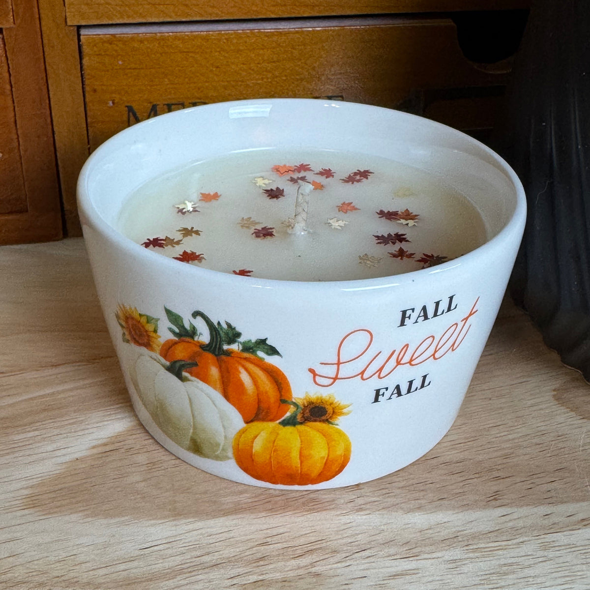 A white ceramic candle jar with a fall-themed design featuring orange and white pumpkins, sunflowers, and the text 'FALL Sweet FALL' on a light wood surface.